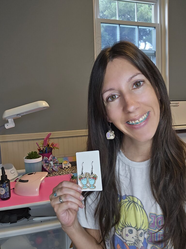 Dana sitting and smiling, holding up a pair of handmade earrings.