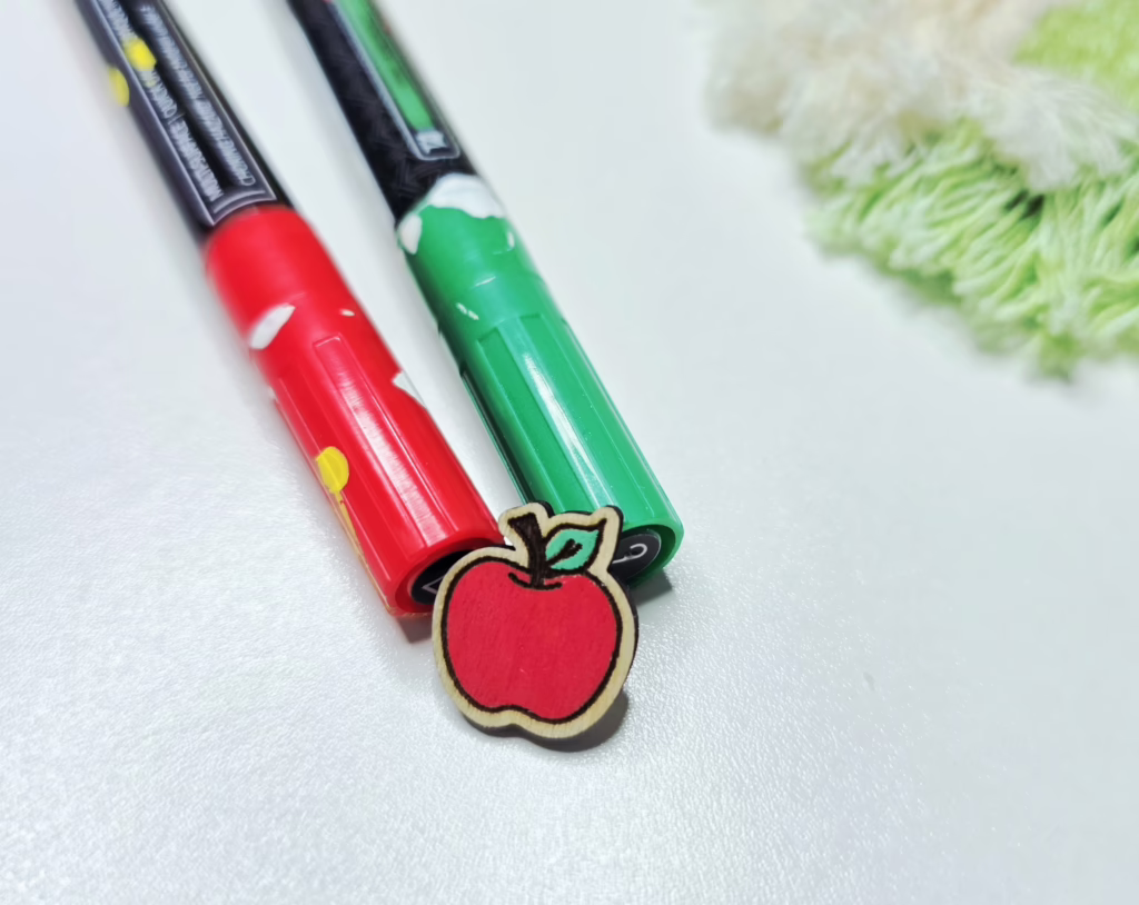 a wooden earring shaped like an apple, sitting in front of green and red markers