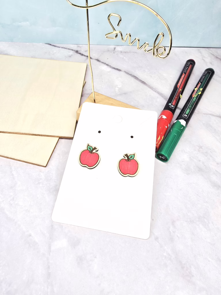 Laser cut apple earrings made from basswood, displayed on an earring card with red and green paint markers beside them.”